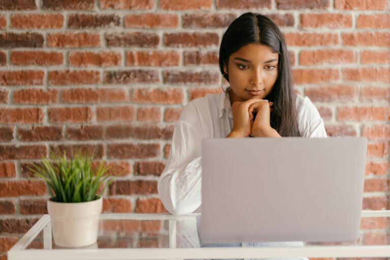 Young woman deeply focused on her laptop studying indoors against a brick wall backdrop.